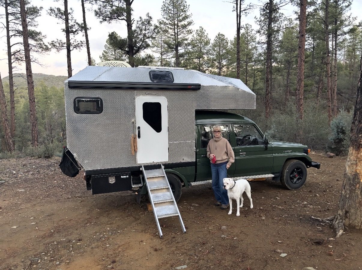 KENDL parked at a forest campsite among tall ponderosa pines, with its ladder down and a woman and white dog standing beside the Land Cruiser