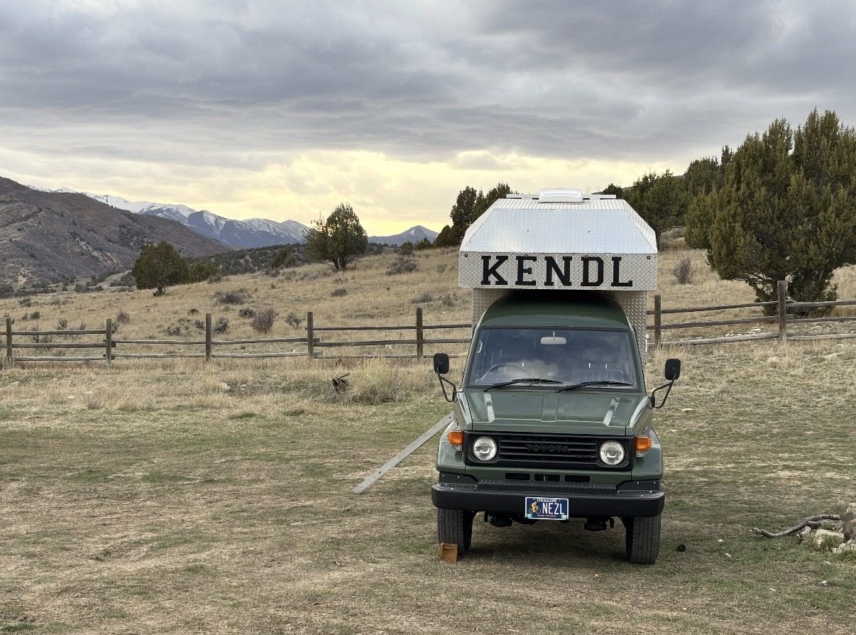 Front view of KENDL on its green right-hand-drive Land Cruiser, parked in a fenced field with snowy mountains in the distance
