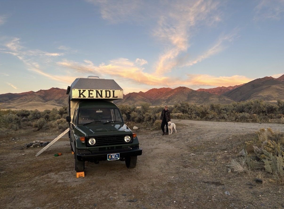 KENDL, a hand-built aluminum diamond-plate camper on a green Toyota Land Cruiser, parked on a gravel road at sunset with red-lit mountains behind